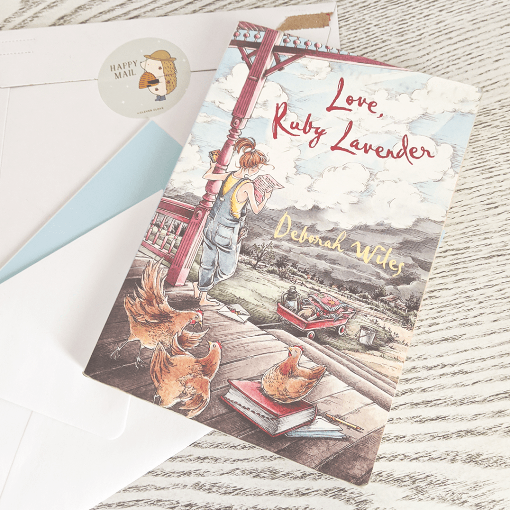 Bookstagram photo featuring LOVE, RUBY LAVENDER by Deborah Wiles. The book rests atop a pile of letters and a package sealed with a sticker. The sticker reads "Happy Mail" and depicts a small cartoon hedgehog wearing a beekeeper's hat and holding a bee hive in gloved hands.