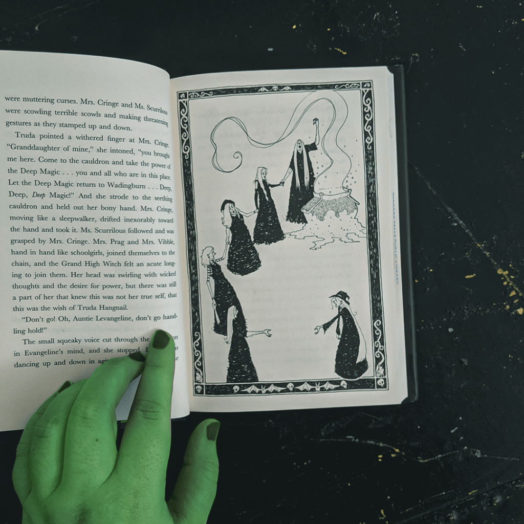 Bookstagram photo featuring an interior illustration from The Bag of Bones by Vivian French. Elza's hand, which has been painted green, holds the book open. The interior illustration features six witches gathered around a steamy cauldron.