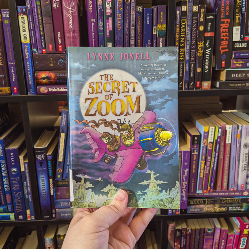 Bookstagram photo features THE SECRET OF ZOOM by Lynne Jonell. Elza holds the boo up against a rainbow-organized shelf of blue, purple, black, and pink books.