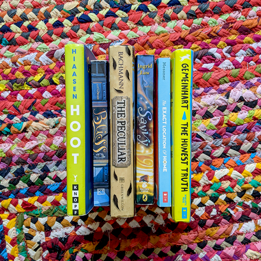Bookstagram photo features six Middle Grade books stacked spine-up on a colorful braided carpet, including HOOT by Carl Hiaasen; BLISS by Kathryn Littlewood; THE PECULIAR by Stefan Bachmann; SAVVY by Ingrid Law; THE EXACT LOCATION OF HOME by Kate Messner; and THE HONEST TRUTH by Dan Gemeinhart