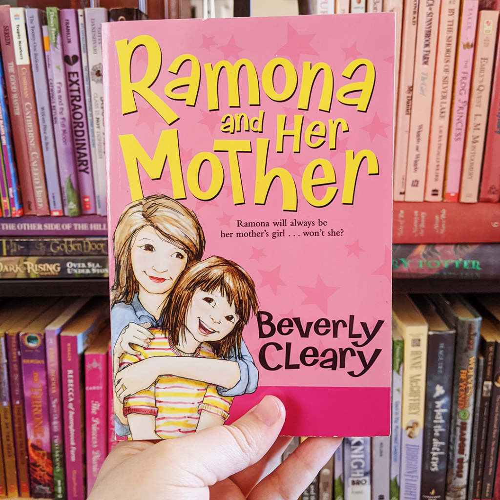 Bookstagram photo featuring RAMONA AND HER MOTHER by Beverly Cleary. Elza holds the book up. In the background, you can see a shelf with pink and purple books.