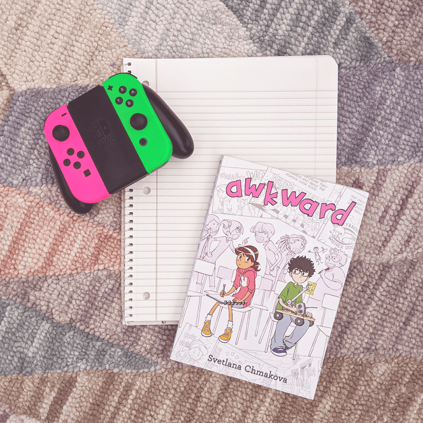 Bookstagram photo featuring AWKWARD by Svetlana Chmakova. The book is placed atop a spiral notebook beside a Nintendo Switch controller.