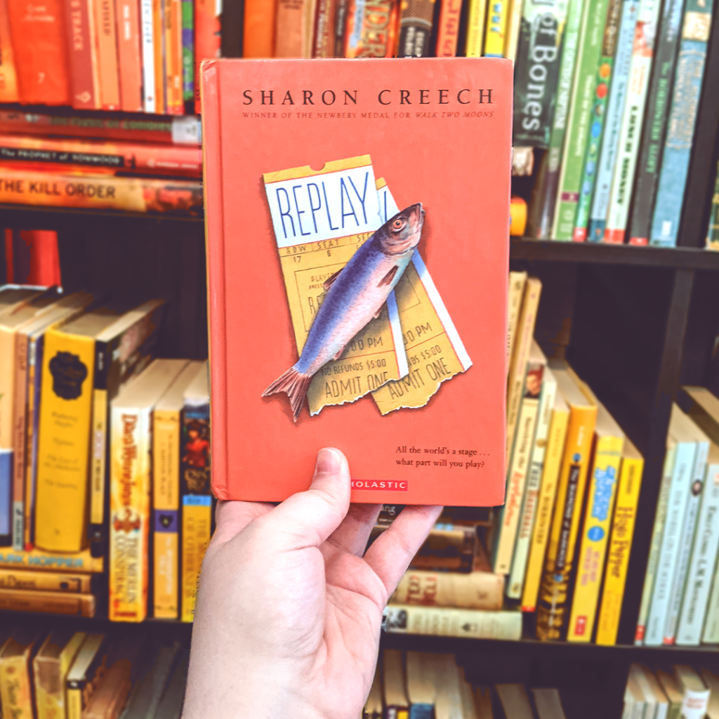 Photograph features the book REPLAY by Sharon Creech. Elza holds the book up. In the background you can see a bookshelf arranged in rainbow order in shades of orange and yellow.