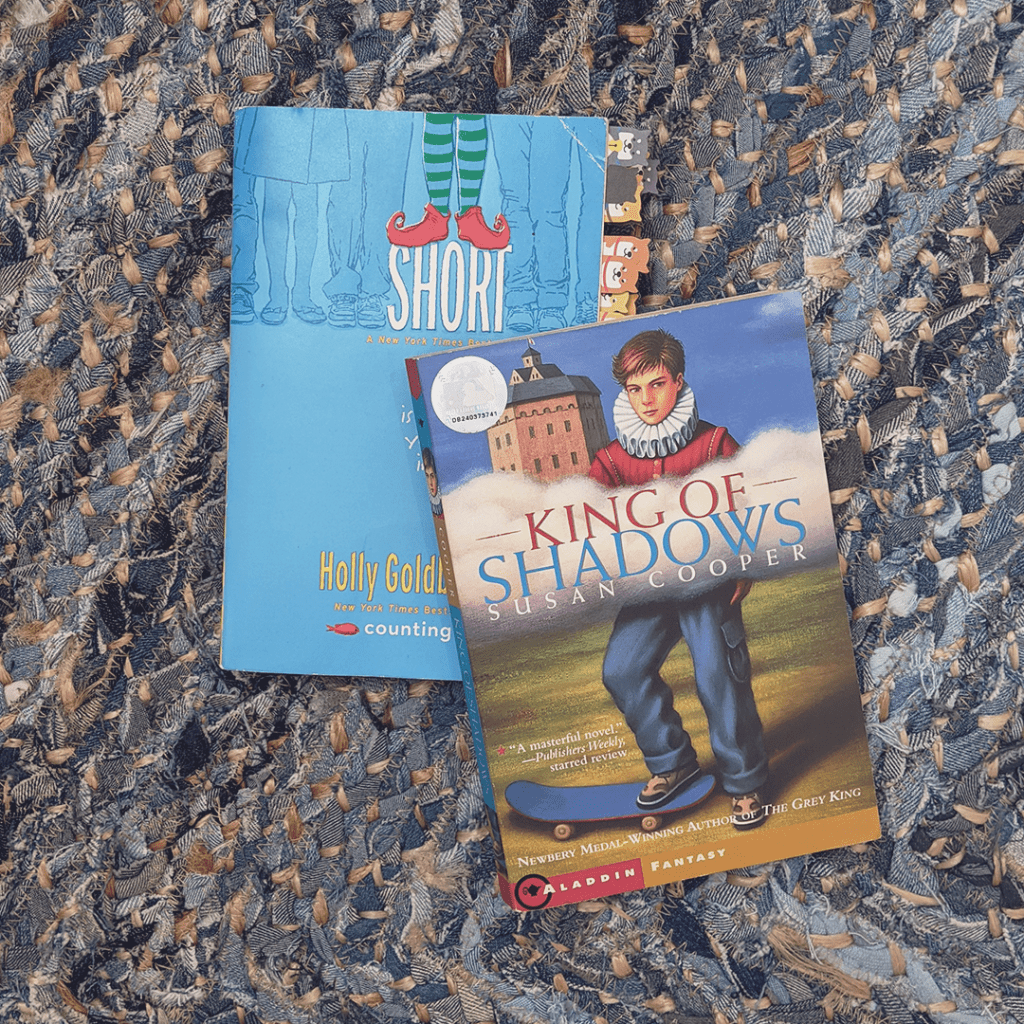 Photograph features the books SHORT by Holly Goldberg Sloan and KING OF SHADOWS by Susan Cooper. They lay on a denim-colored braided rug.